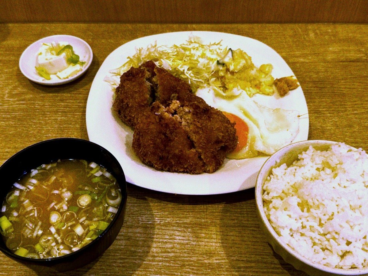 A minced meat cutlet set meal with rice, miso soup, tofu, and shredded cabbage, served on a wooden table.